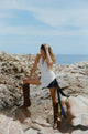 Woman sitting on rocks by the ocean wearing a white wrap top and brown boots.