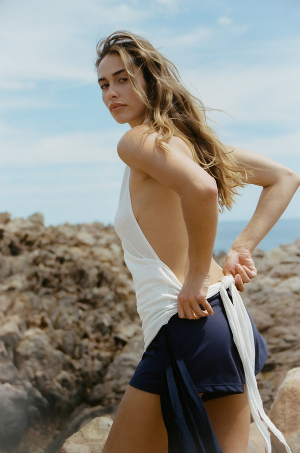 Woman in a white low back top and navy shorts standing on rocky terrain with a clear sky.