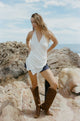 Woman in a white sleeveless wrap top and brown boots standing on rocky terrain with ocean in the background